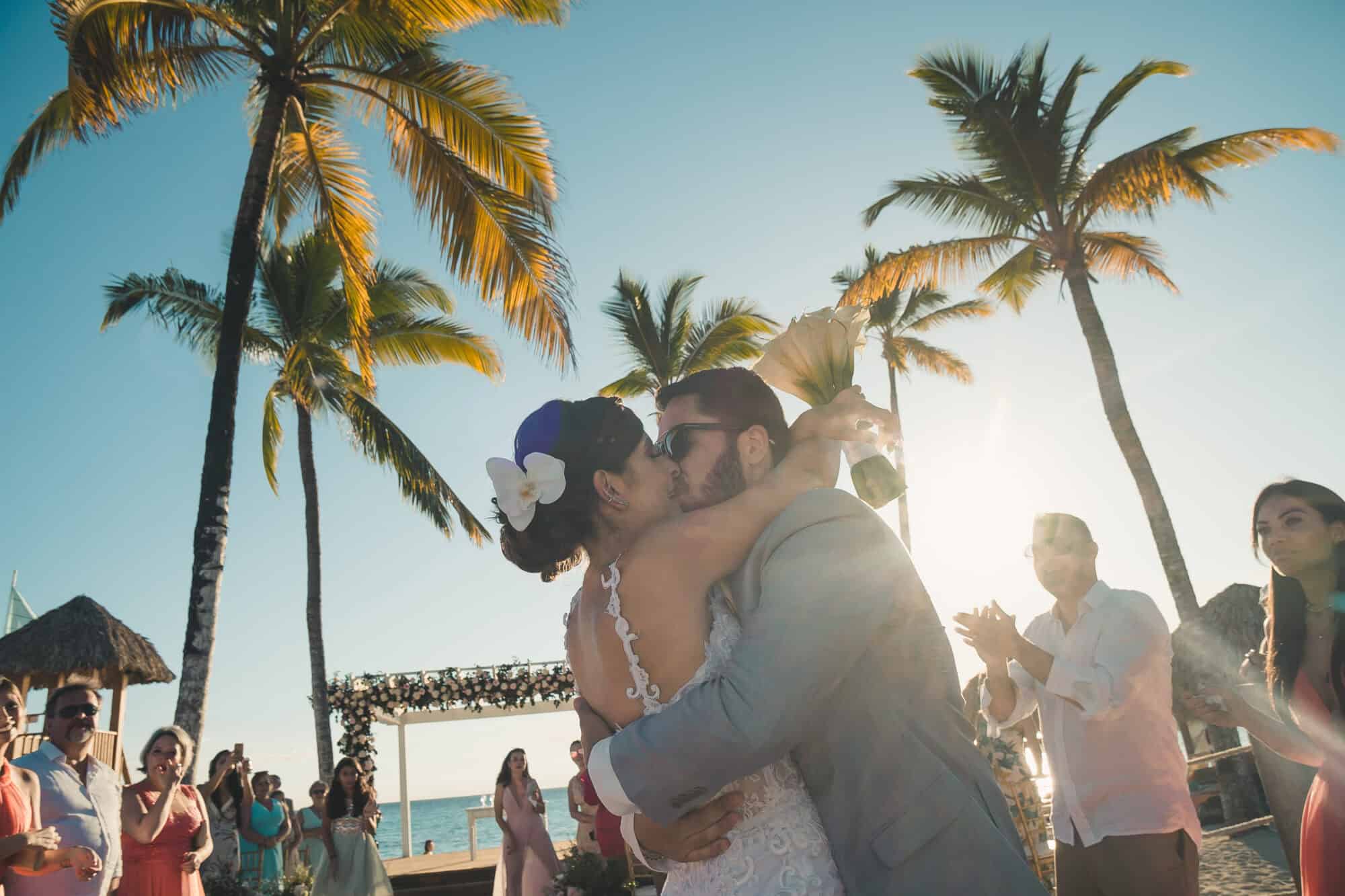 Fotografia mostra casal celebrando o casamento no Caribe cercado de convidados, céu azul e palmeiras