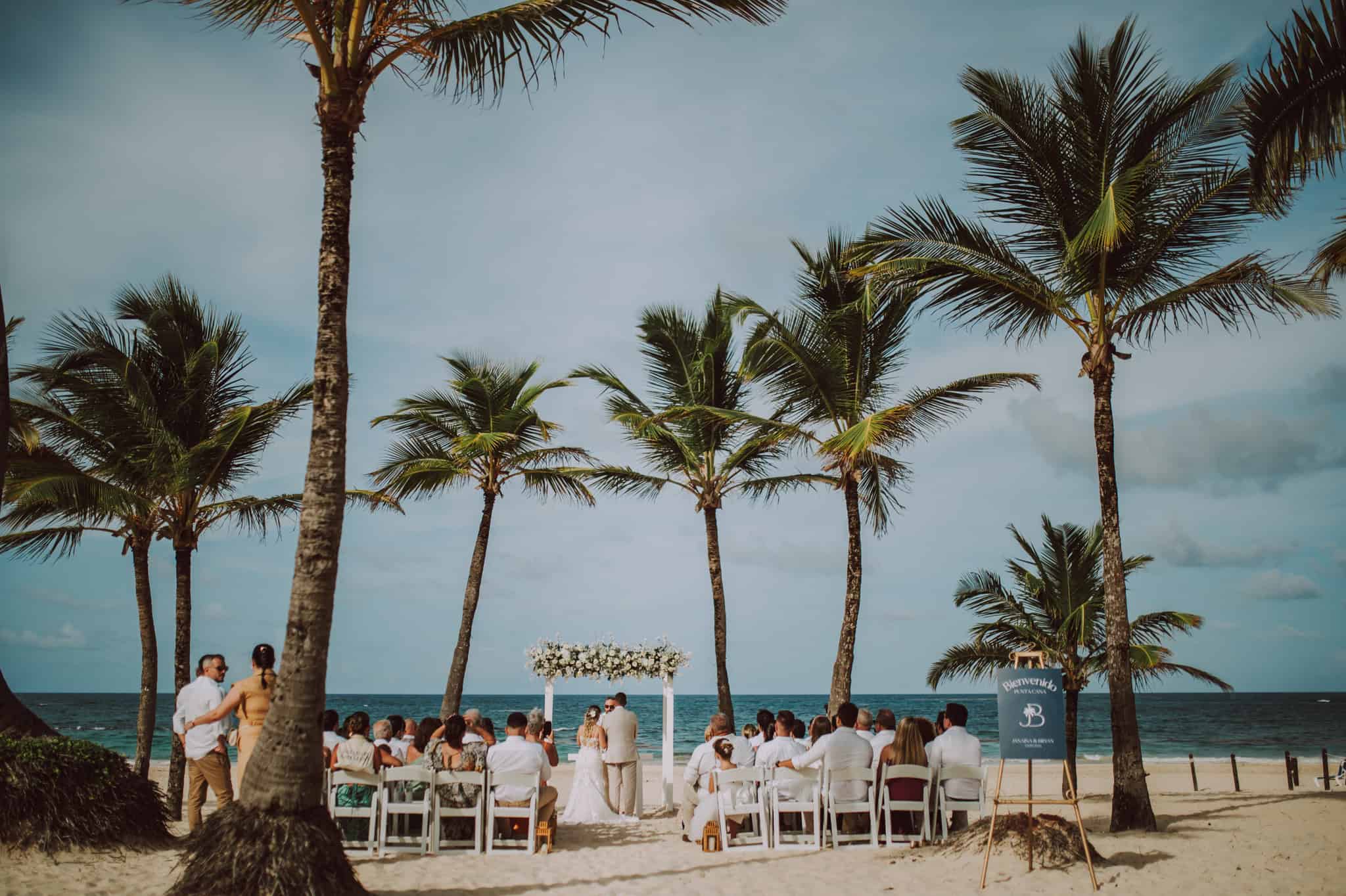 Cerimônia de casamento na praia com arco floral, convidados sentados e mar ao fundo em destination wedding.