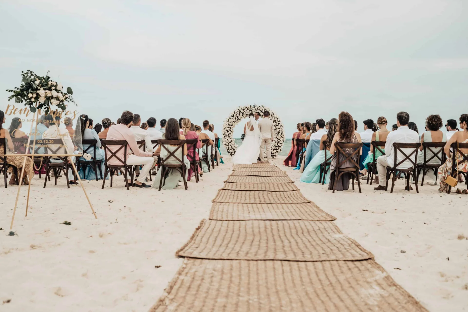 Corredor da cerimônia na praia, com os convidados sentados e o casal sob o arco redondo de flores, de frente para o mar