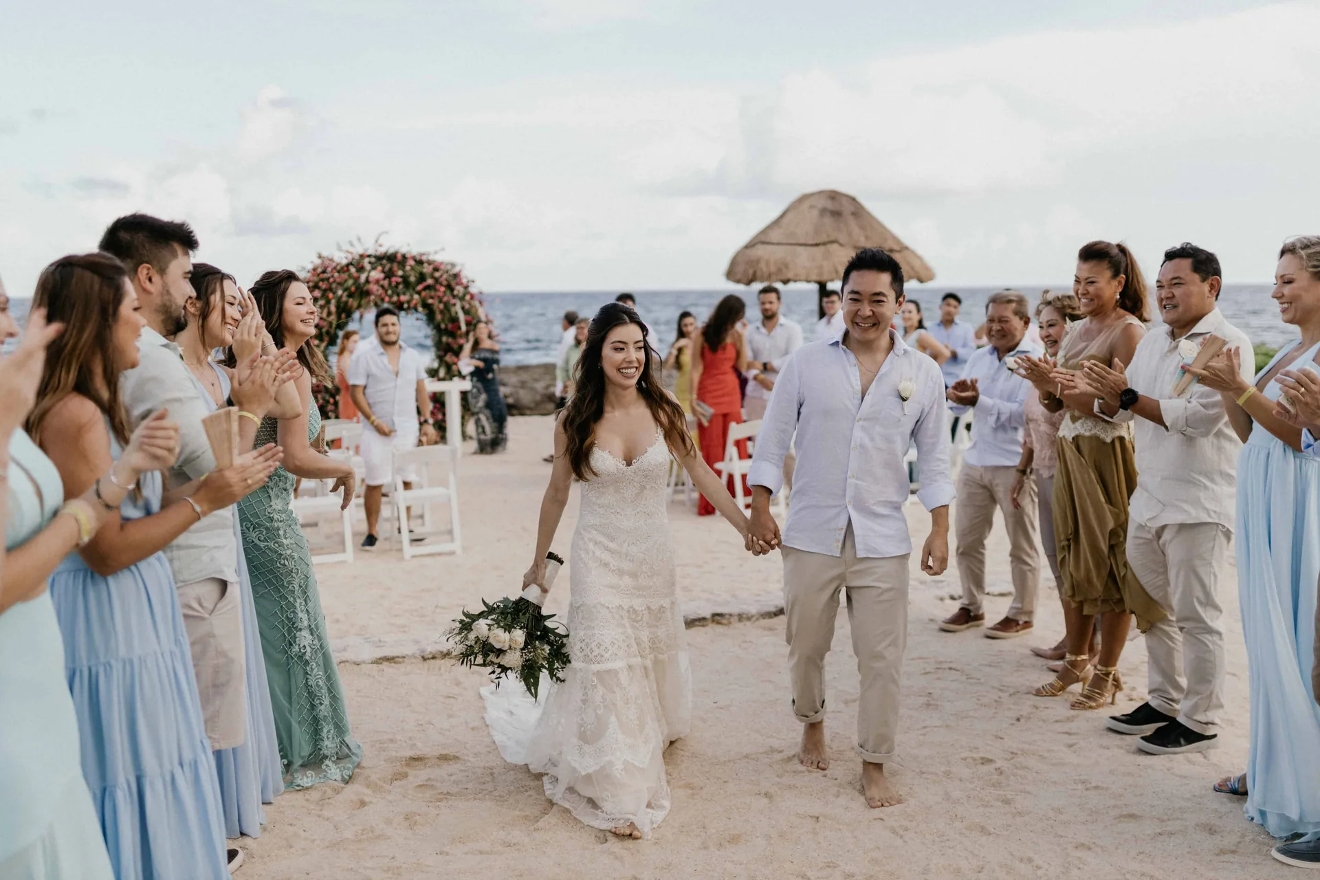 Casal caminhando descalço na areia entre os convidados que aplaudem, com o mar ao fundo