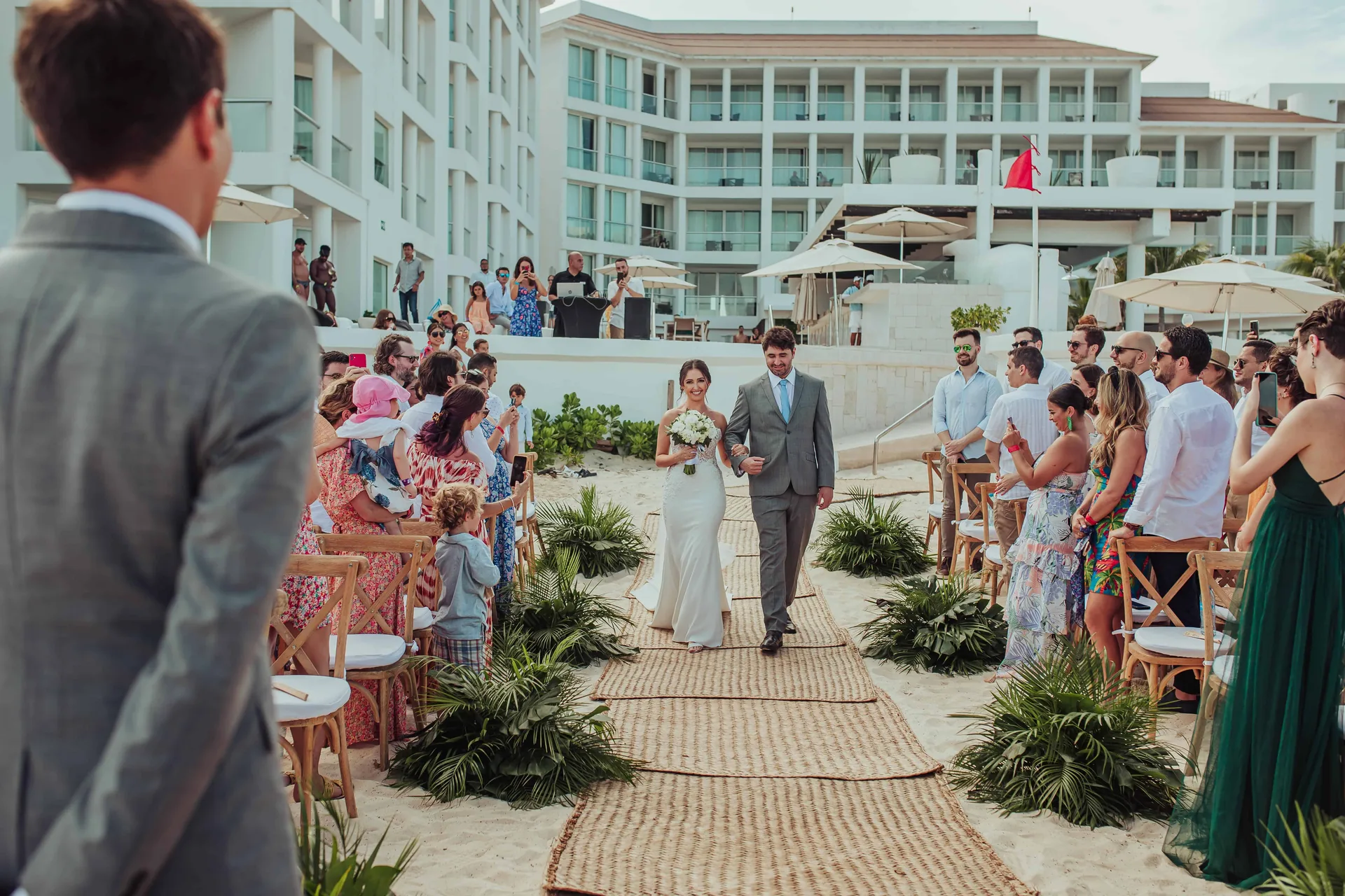 Casal entrando na cerimônia montada na areia, com os convidados sentados e o resort ao fundo