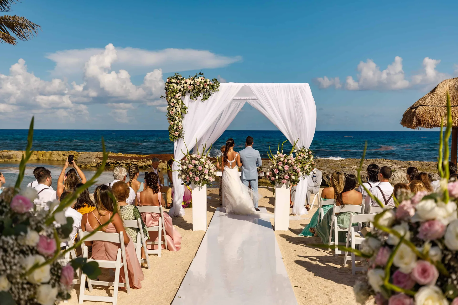 Cerimônia montada com tecido branco e flores diante da praia, com a costa rochosa ao fundo