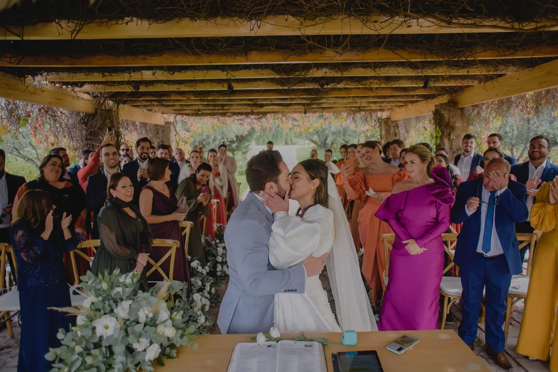Casal se beijando sob o pergolado de parreiras, com os convidados em pé aplaudindo em volta