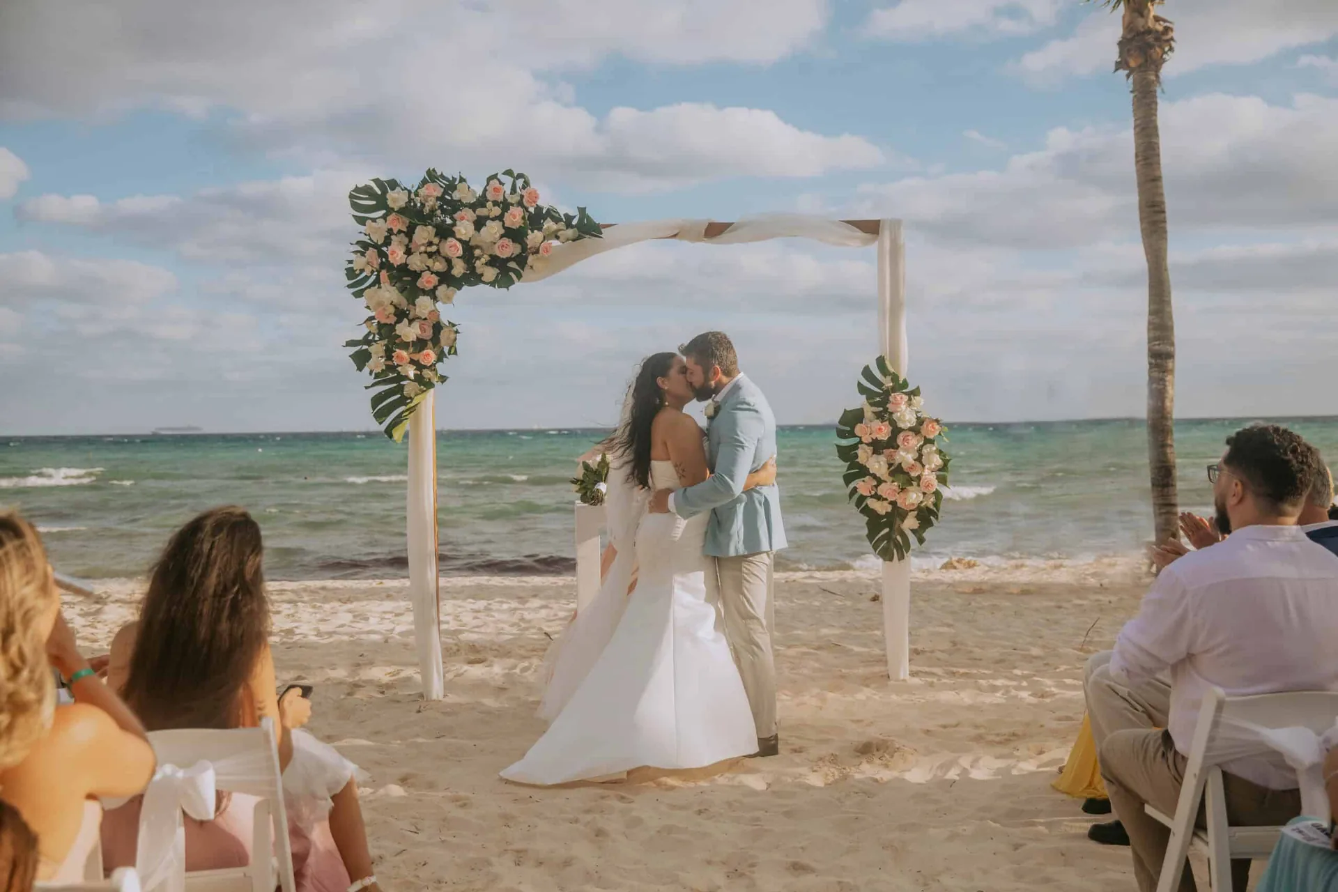 Casal se beijando sob o arco de flores montado na areia, com o mar turquesa atrás e os convidados nos dois lados