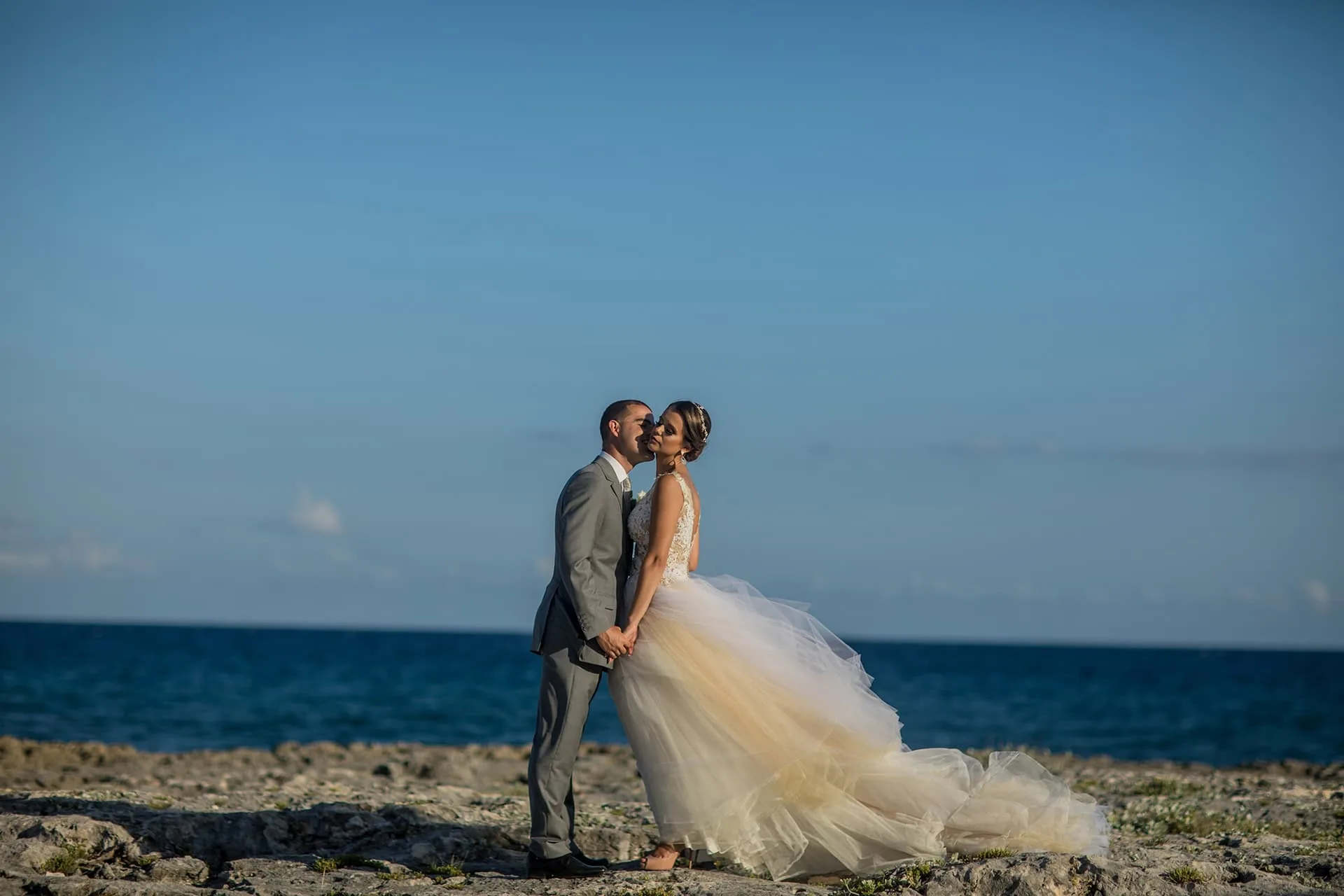 Casal abraçado na costa rochosa, com o mar azul atrás, no fim da tarde