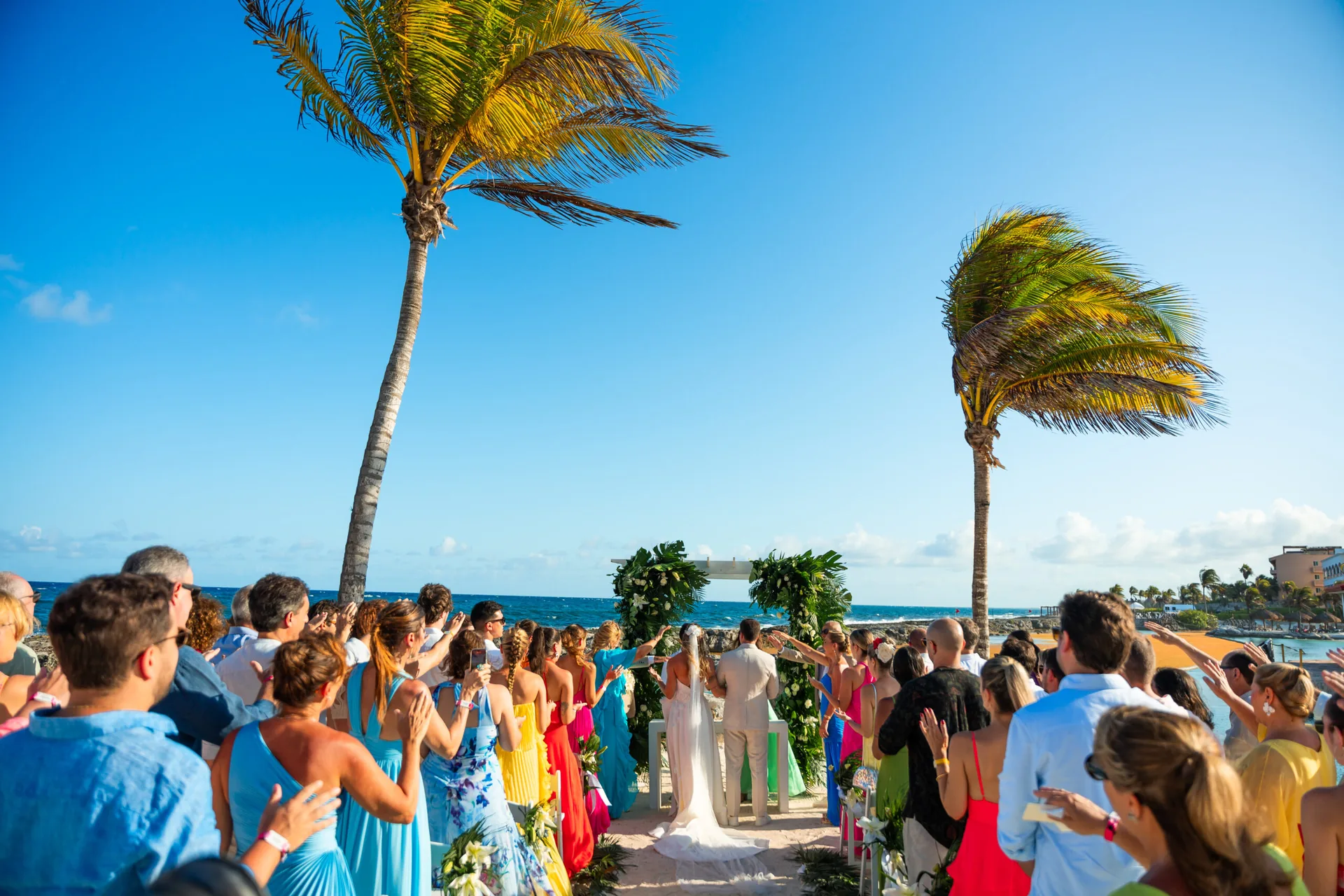 Cerimônia entre coqueiros, com os convidados em pé aplaudindo o casal diante do mar do Caribe