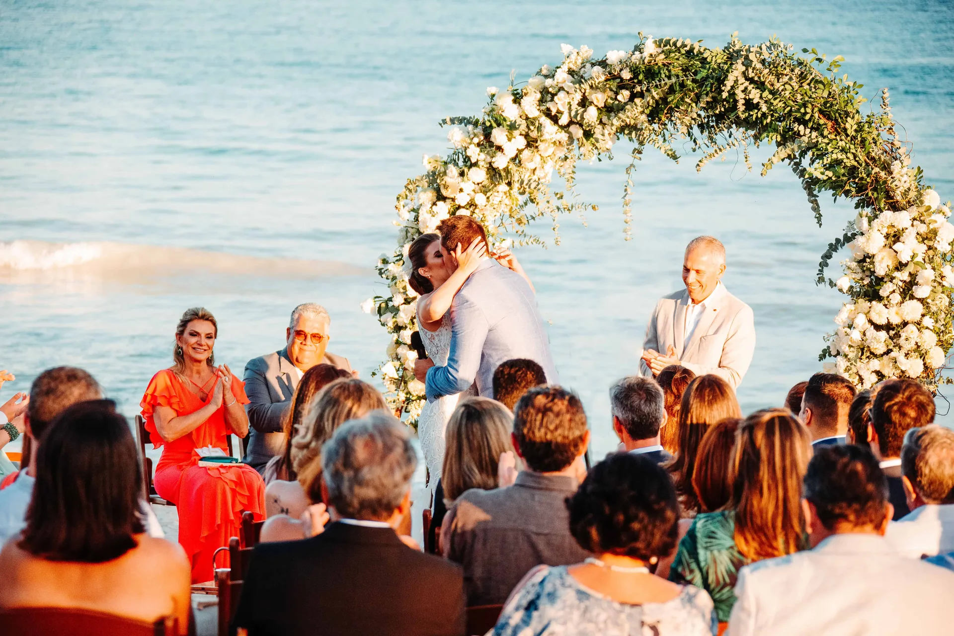Cerimônia na praia sob um arco redondo de flores, com os convidados sentados de frente para o mar