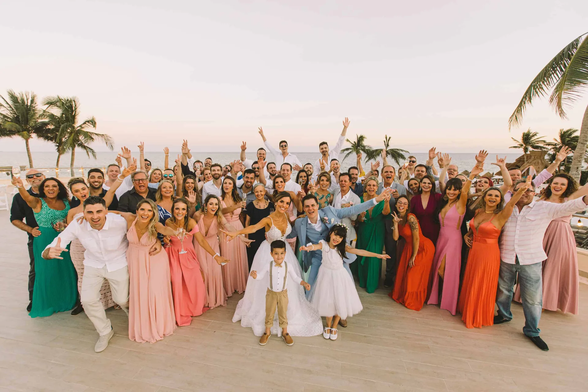 Foto de grupo com todos os convidados de braços erguidos ao lado do casal, com coqueiros e o mar ao fundo