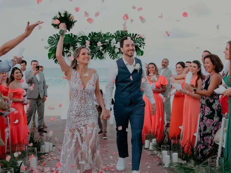 Casal de mãos dadas saindo da cerimônia sob chuva de pétalas, com o mar do Caribe ao fundo