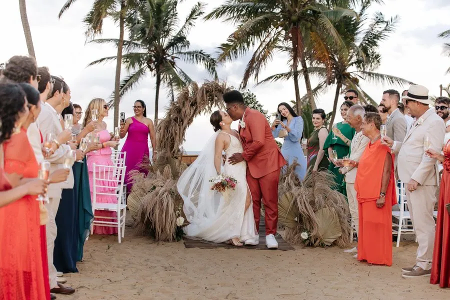 Casal se beijando na saída da cerimônia na praia, brindado pelos convidados