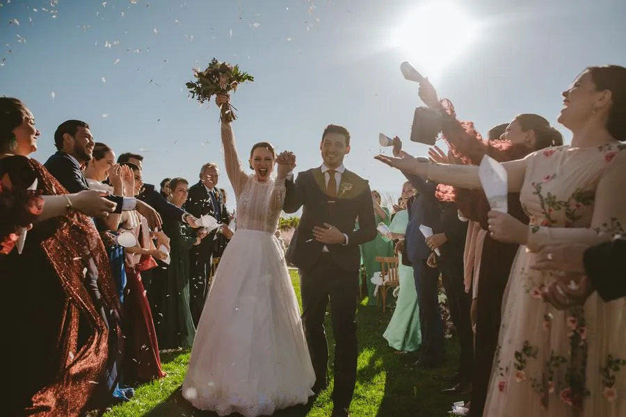 Casal saindo da cerimônia sob chuva de pétalas, entre os convidados, contra o sol